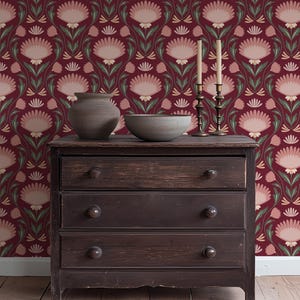 May include: A dark wooden dresser with three drawers, topped with a gray ceramic vase, a gray bowl, and two brass candle holders with lit candles. The background features a burgundy floral wallpaper with pink and green accents.