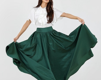 May include: A woman wearing a white blouse with a ruffled collar and a long, flowing green skirt. She is standing with her arms outstretched, holding the skirt out to the sides.