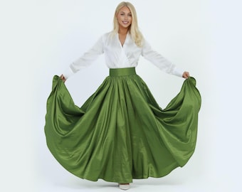May include: A long, flowing green skirt with a wide waistband is paired with a white wrap-style blouse. The skirt's fabric has a subtle sheen, and the outfit is set against a plain white background. The skirt appears to be floor-length.