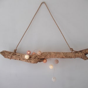 Puede incluir: Una rama de madera natural con una cadena de luces de bolas de algodón blancas y rosas colgando de ella. La rama está suspendida del techo con una cuerda.