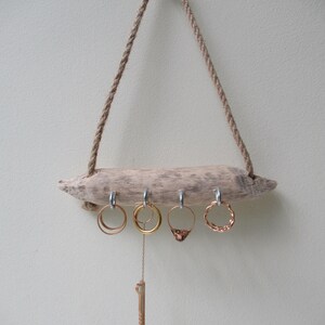 May include: A rustic wooden jewelry holder with a rope hanger and five metal hooks. The holder is decorated with gold rings and a gold chain.