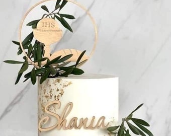 May include: White cake with gold flecks and a wooden topper with a chalice and the letters "IHS" and the name "Shania". The cake is decorated with green leaves and blueberries.
