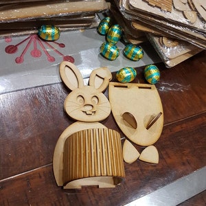 May include: A wooden bunny rabbit Easter basket kit with a cylindrical base and a separate bunny head and ears. The kit is disassembled and ready to be assembled. There are also several chocolate Easter eggs wrapped in gold and turquoise foil.