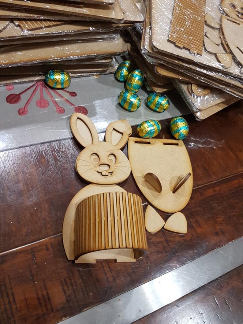 May include: A wooden bunny rabbit Easter basket kit with a cylindrical base and a separate bunny head and ears. The kit is disassembled and ready to be assembled. There are also several chocolate Easter eggs wrapped in gold and turquoise foil.