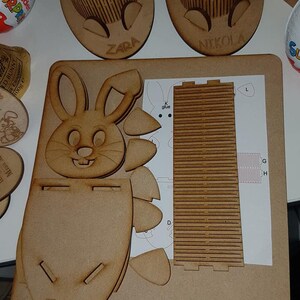 May include: A wooden Easter bunny craft kit with the name "ZARA" engraved on the front. The kit includes a bunny shaped box, a separate piece for the back of the box, and a sheet of instructions with the letters "G", "H", "L", and "K glue" printed on it.