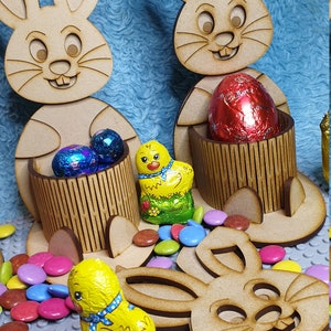 May include: Three wooden bunny rabbit Easter egg holders with a chocolate chick and chocolate eggs. The bunnies are holding the eggs in their paws. The bunnies are brown and have white ears. The chocolate chick is yellow and has a blue ribbon around its neck. The chocolate eggs are wrapped in foil and are red, blue, and silver.
