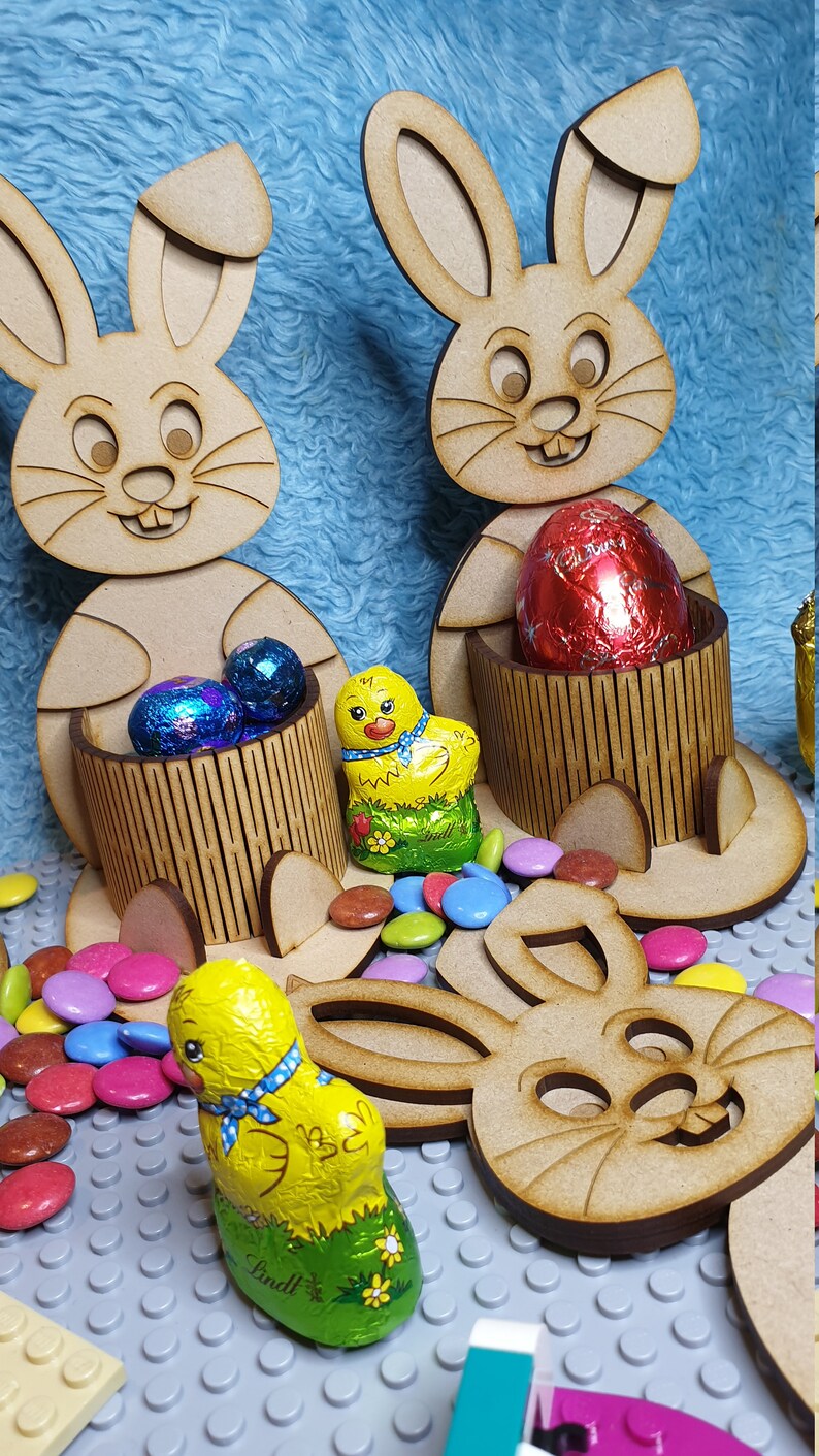 May include: Three wooden bunny rabbit Easter egg holders with a chocolate chick and chocolate eggs. The bunnies are holding the eggs in their paws. The bunnies are brown and have white ears. The chocolate chick is yellow and has a blue ribbon around its neck. The chocolate eggs are wrapped in foil and are red, blue, and silver.