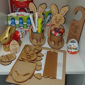 May include: A wooden Easter bunny craft kit with a bunny-shaped stand, a bunny-shaped holder, and a bunny-shaped door hanger. The kit includes a sheet of instructions, a sheet of wood pieces, and a sheet of paper with a grid pattern. The kit also includes a Kinder Surprise egg and a small bag of chocolate eggs.