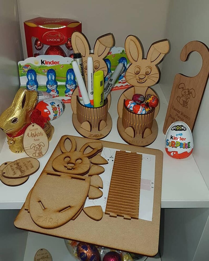 May include: A wooden Easter bunny craft kit with a bunny-shaped stand, a bunny-shaped holder, and a bunny-shaped door hanger. The kit includes a sheet of instructions, a sheet of wood pieces, and a sheet of paper with a grid pattern. The kit also includes a Kinder Surprise egg and a small bag of chocolate eggs.