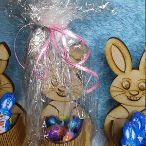 May include: Wooden bunny-shaped Easter basket with a pink ribbon and cellophane wrapping. The basket is filled with colorful chocolate eggs. The basket is labeled "ZARA".