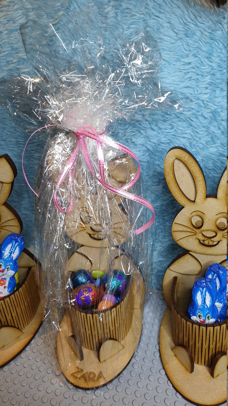 May include: Wooden bunny-shaped Easter basket with a pink ribbon and cellophane wrapping. The basket is filled with colorful chocolate eggs. The basket is labeled "ZARA".