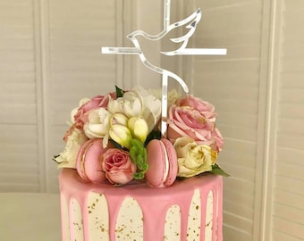 May include: A cake with pink frosting and gold flecks, topped with a silver dove and cross cake topper. The cake is decorated with pink macarons and fresh flowers, including roses and white blossoms. A celebratory dessert.