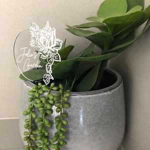 May include: A white heart-shaped acrylic plant marker with the text "Julie's Garden" and a stylised rose design. The marker is inserted into a grey ceramic pot with a succulent plant.
