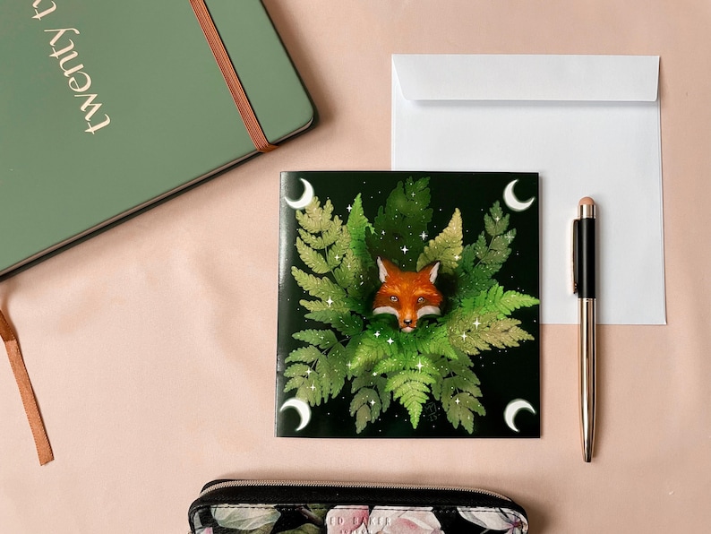Illustrated greeting card Fox in Ferns.
Square card picturing a fox's head peeking out of ferns. Dark green background, with a crescent moon in each corner.
The photo also shows a white envelope that is included.