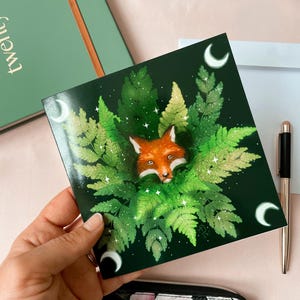 Square card picturing a fox's head peeking out of ferns. Dark green background, with a crescent moon in each corner.