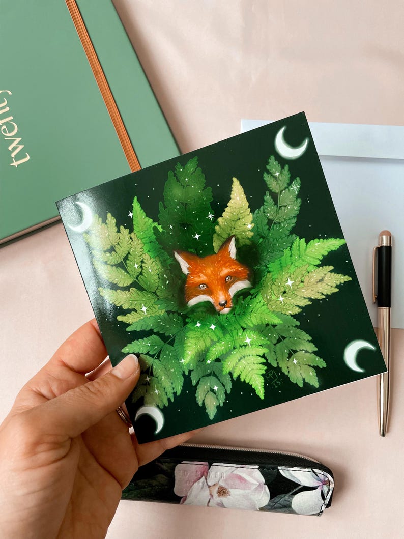 Square card picturing a fox's head peeking out of ferns. Dark green background, with a crescent moon in each corner.