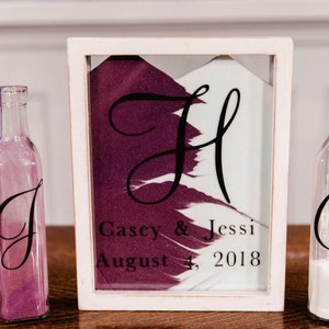 May include: A wedding sand ceremony set. A framed sand art piece displays the initials "J" and "H", the names "Casey & Jessi", and the date "August 4, 2018". Two glass bottles, one with "J" and purple sand, the other with "C" and white sand.