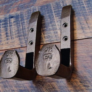 May include: Two rustic metal hooks with a dark brown finish. Each hook has a rounded top with the letters "DH" and "L" engraved on it. The hooks are mounted on a wooden surface.