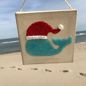 Puede incluir: Un peque&ntilde;o lienzo blanco con una ballena azul que lleva un gorro de Pap&aacute; Noel rojo. La ballena est&aacute; hecha de purpurina azul y el gorro de purpurina roja. El lienzo est&aacute; colgado de una cuerda.