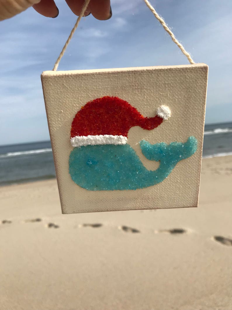 Puede incluir: Un peque&ntilde;o lienzo blanco con una ballena azul que lleva un gorro de Pap&aacute; Noel rojo. La ballena est&aacute; hecha de purpurina azul y el gorro de purpurina roja. El lienzo est&aacute; colgado de una cuerda.