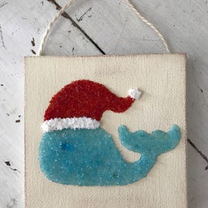 Puede incluir: Una ballena azul con un gorro de Pap&aacute; Noel rojo con ribete blanco, hecha de purpurina y pegada a un lienzo blanco. El lienzo tiene una cuerda para colgar.