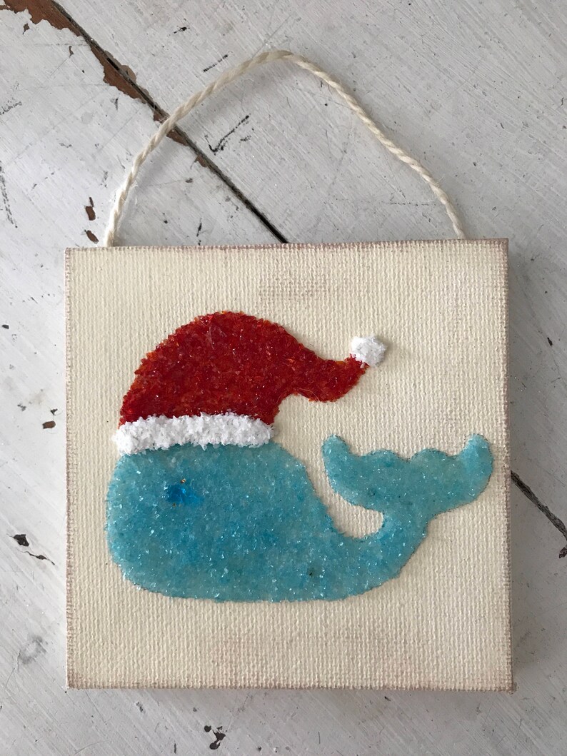 Puede incluir: Una ballena azul con un gorro de Pap&aacute; Noel rojo con ribete blanco, hecha de purpurina y pegada a un lienzo blanco. El lienzo tiene una cuerda para colgar.