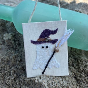 Puede incluir: Un peque&ntilde;o lienzo blanco con un fantasma que lleva un sombrero de bruja morado y sostiene una escoba hecha de hilo blanco y dorado. El fantasma tiene dos ojos negros y una concha para la nariz. El lienzo est&aacute; colgado de una cuerda.