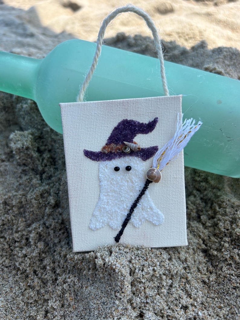 Puede incluir: Un peque&ntilde;o lienzo blanco con un fantasma que lleva un sombrero de bruja morado y sostiene una escoba hecha de hilo blanco y dorado. El fantasma tiene dos ojos negros y una concha para la nariz. El lienzo est&aacute; colgado de una cuerda.