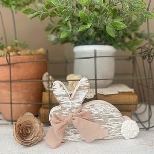 May include: Whitewashed wooden bunny rabbit figurine with a pink bow. The bunny is sitting on a white surface with a brown paper rose and a white fluffy tail.