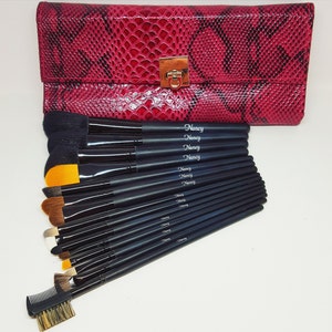 May include: A set of 14 black makeup brushes with gold accents, arranged in a fan shape. The brushes are in a pink snakeskin-patterned case with a gold clasp. The brushes are labeled "Nancy" on the handles.