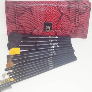 May include: A set of black makeup brushes with yellow and brown bristles, arranged in a row. The brushes are labeled "Oprah" and come in a red snakeskin-patterned case.