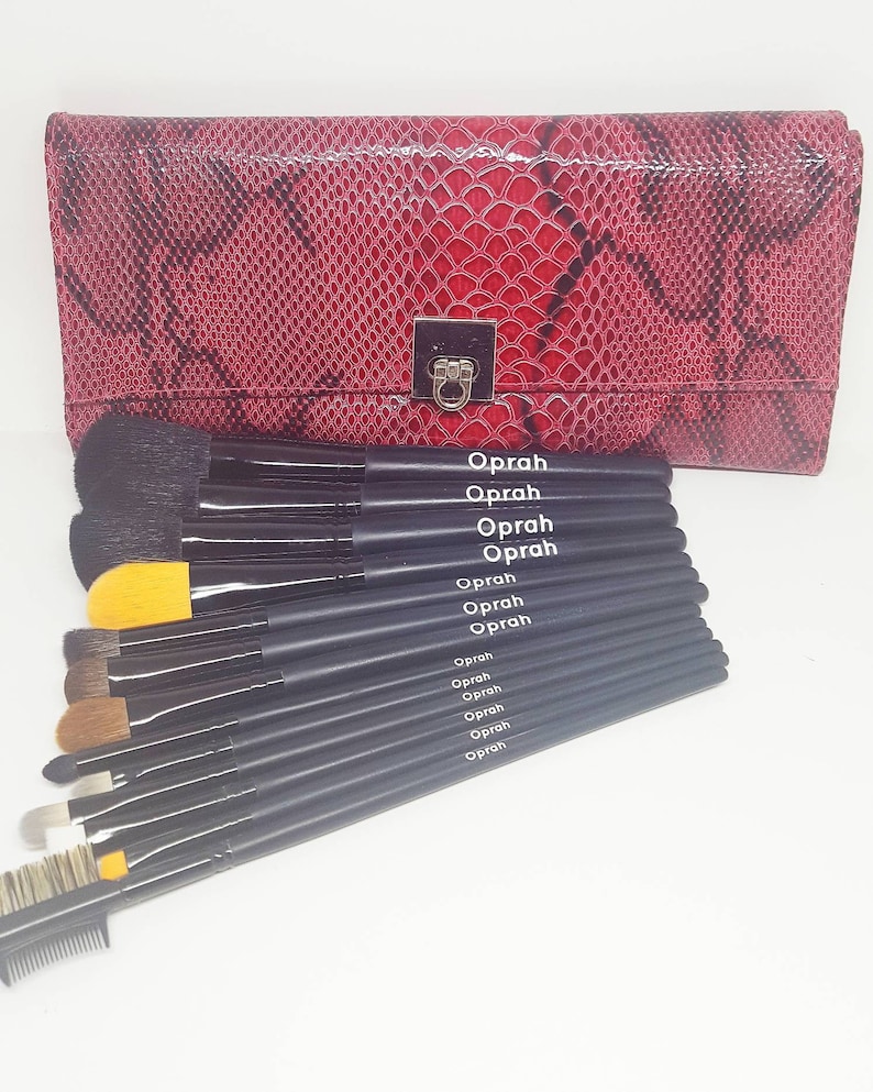 May include: A set of black makeup brushes with yellow and brown bristles, arranged in a row. The brushes are labeled "Oprah" and come in a red snakeskin-patterned case.