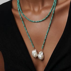 May include: A double-strand necklace with turquoise beads and two large, irregular white pearls. The necklace has a gold clasp and is set against a black backdrop.