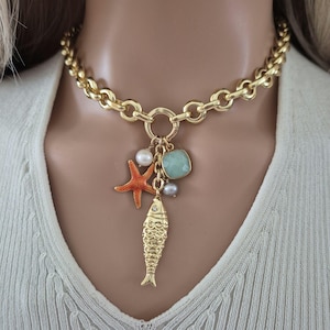 Könnte beinhalten: Goldkette mit einem Anhänger in Form eines goldenen Fisches, eines roten Seesterns, eines hellgrünen Edelsteins und weißen Perlen.