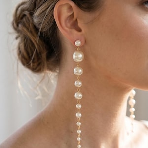 May include: Elegant, long pearl earrings. The earrings feature a small pearl stud at the top, followed by a series of graduated, round pearls suspended on a delicate gold chain. The pearls vary in size, creating a cascading effect.