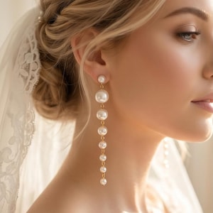 May include: Elegant gold earrings with graduated white pearls. The earrings dangle from the earlobe, with a small pearl at the top and larger pearls cascading down. The earrings are a classic bridal accessory.