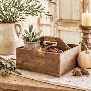 Könnte beinhalten: Ein rechteckiger Holzkasten mit einem mittigen Griff, der mit Jutegarn umwickelt ist. Der Kasten verfügt über mehrere Fächer, die eine kleine Topfpflanze, ein Glas-Teelichthalter mit brennender Kerze und mehrere braune Eicheln enthalten.