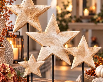 Puede incluir: Cuatro estrellas de cinco puntas con un acabado beige claro texturizado, cada una montada sobre una varilla de metal negro delgada. Las estrellas varían en tamaño y están dispuestas a diferentes alturas, creando una exhibición de adornos en capas.