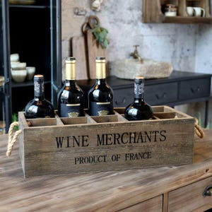 Könnte beinhalten: Eine rechteckige Holzkiste mit Seilgriffen, die den schwarzen Schriftzug WINE MERCHANTS PRODUCT OF FRANCE trägt. Die Kiste enthält drei dunkle Glasweinflaschen mit goldenen Etiketten, die in einzelnen Fächern innerhalb der Holzkiste stehen.