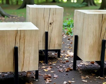 Puede incluir: Tres taburetes cúbicos de madera con vetas naturales y patas de metal negro. Los taburetes están al aire libre en un camino con hojas caídas, rodeados de árboles. La madera tiene nudos y grietas visibles.
