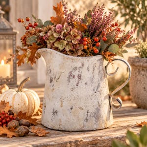 Könnte beinhalten: Ein verwitterter, cremeweißer Keramikkrug mit abgenutzter Oberfläche und geschwungenem Henkel, gefüllt mit einem herbstlichen Arrangement aus getrockneten Hortensienblüten, orangen Beeren, violettem Heidekraut und braunen Ahornblättern.