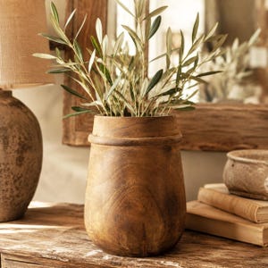 Könnte beinhalten: Eine zylindrische Holzvase mit glatter, dunkelbrauner Oberfläche und einem markanten horizontalen Wulst nahe dem oberen Rand. Die Vase ist mit mehreren grünen Olivenzweigen gefüllt, deren schmale, spitze Blätter nach oben ragen.