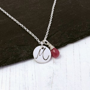 May include: A silver necklace with a small round pendant featuring the letter 'M' and a dangling red gemstone.