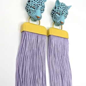 May include: A pair of earrings with blue leopard heads with black spots, yellow details, and long lavender fringe. The earrings have gold-coloured hoops. The background is white.