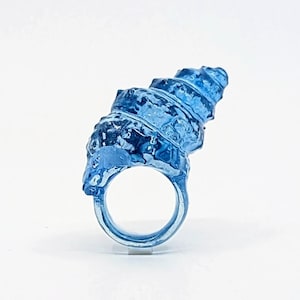 Könnte beinhalten: Ein einzigartiger, hellblauer Ring mit einem strukturierten, muschelartigen Design. Der Ring hat eine spiralförmige Form und einen glatten Ring. Dieser Ring ist ein Statement-Stück, perfekt, um jedem Outfit einen Hauch von künstlerischem Flair zu verleihen.