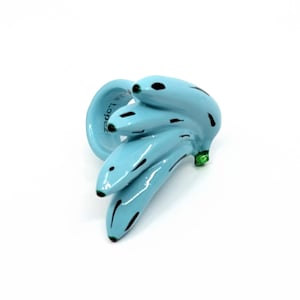 Puede incluir: Un anillo azul claro que presenta un racimo de plátanos con detalles pintados en negro y un pequeño acento verde en la base. El interior de la banda tiene inscrita la palabra 'Lia Lopez' en una fuente oscura, visible sobre el acabado brillante.