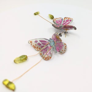 May include: A pair of pink and blue butterfly earrings with gold chains and green glass beads. The butterflies have a white background with pink and orange accents.