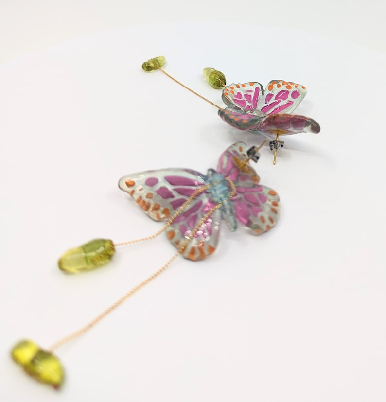 May include: A pair of pink and blue butterfly earrings with gold chains and green glass beads. The butterflies have a white background with pink and orange accents.