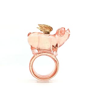 May include: A pink glass ring with a pig figurine on top. The pig has a gold wing on its back.