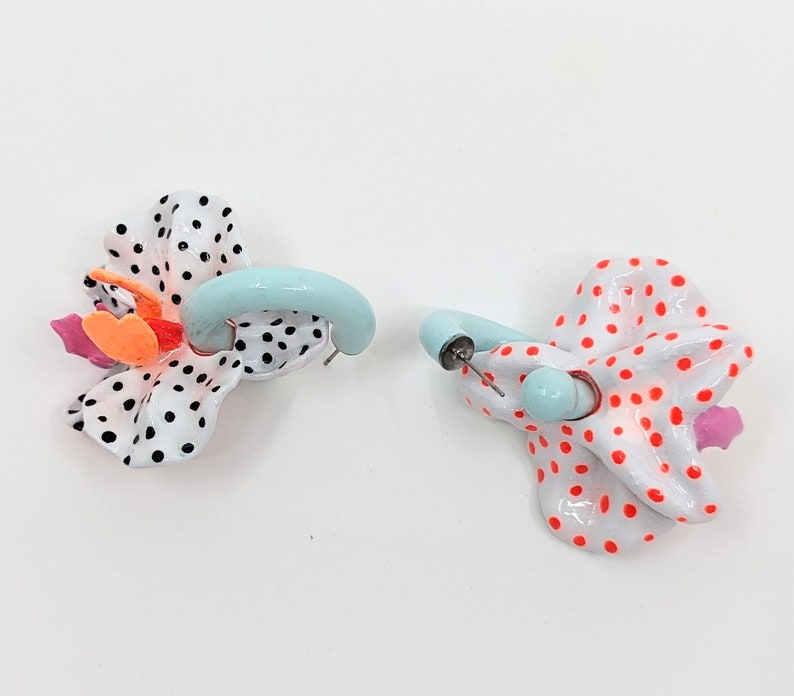 May include: A pair of earrings with a white and black polka dot design and a light blue hoop. One earring has a pink and orange flower accent, while the other has a red and white polka dot design.